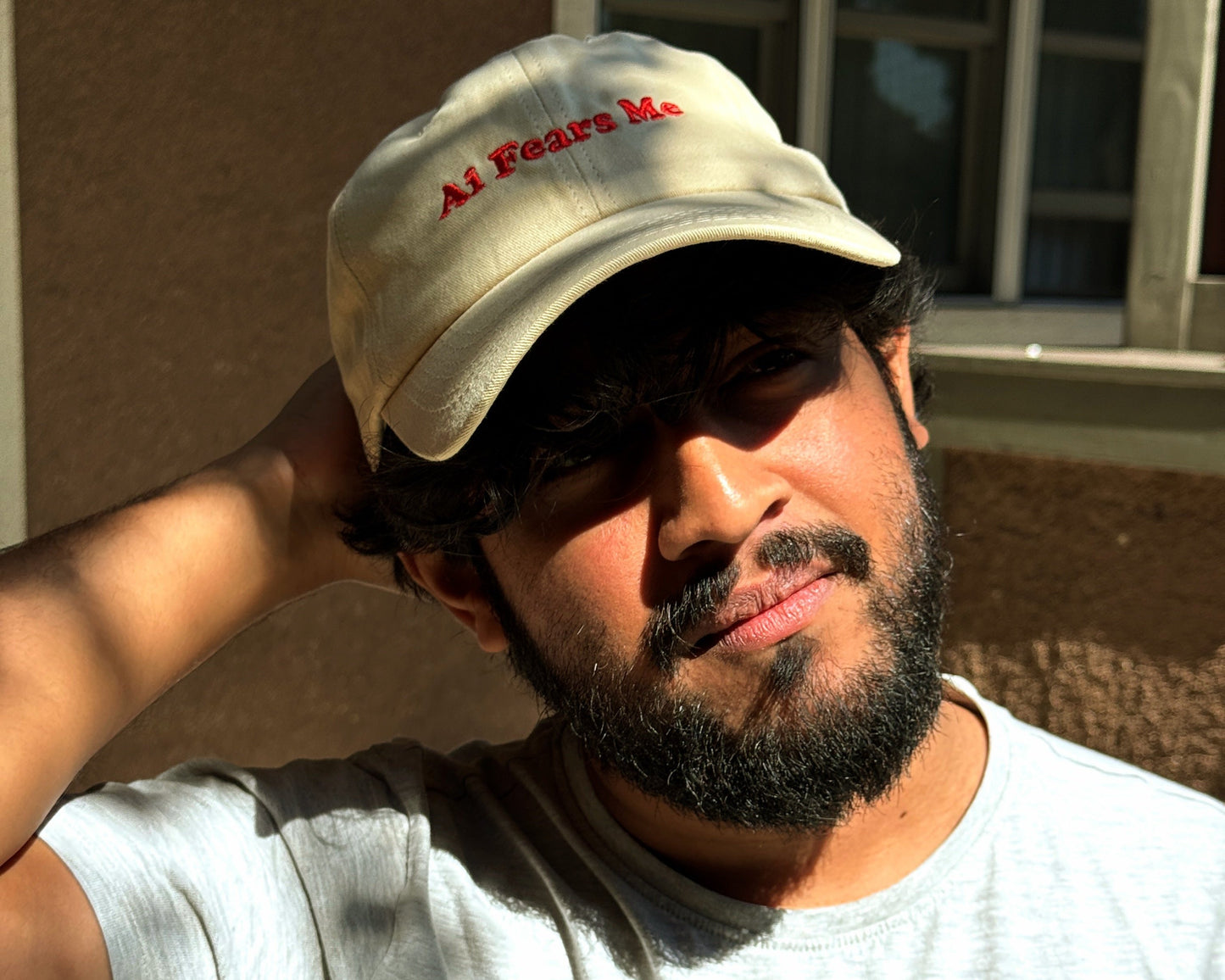 Person wearing a beige dad hat with the words 'AI Fears Me' embroidered in red, standing outdoors in the sunlight with a casual expression and hand resting on their head