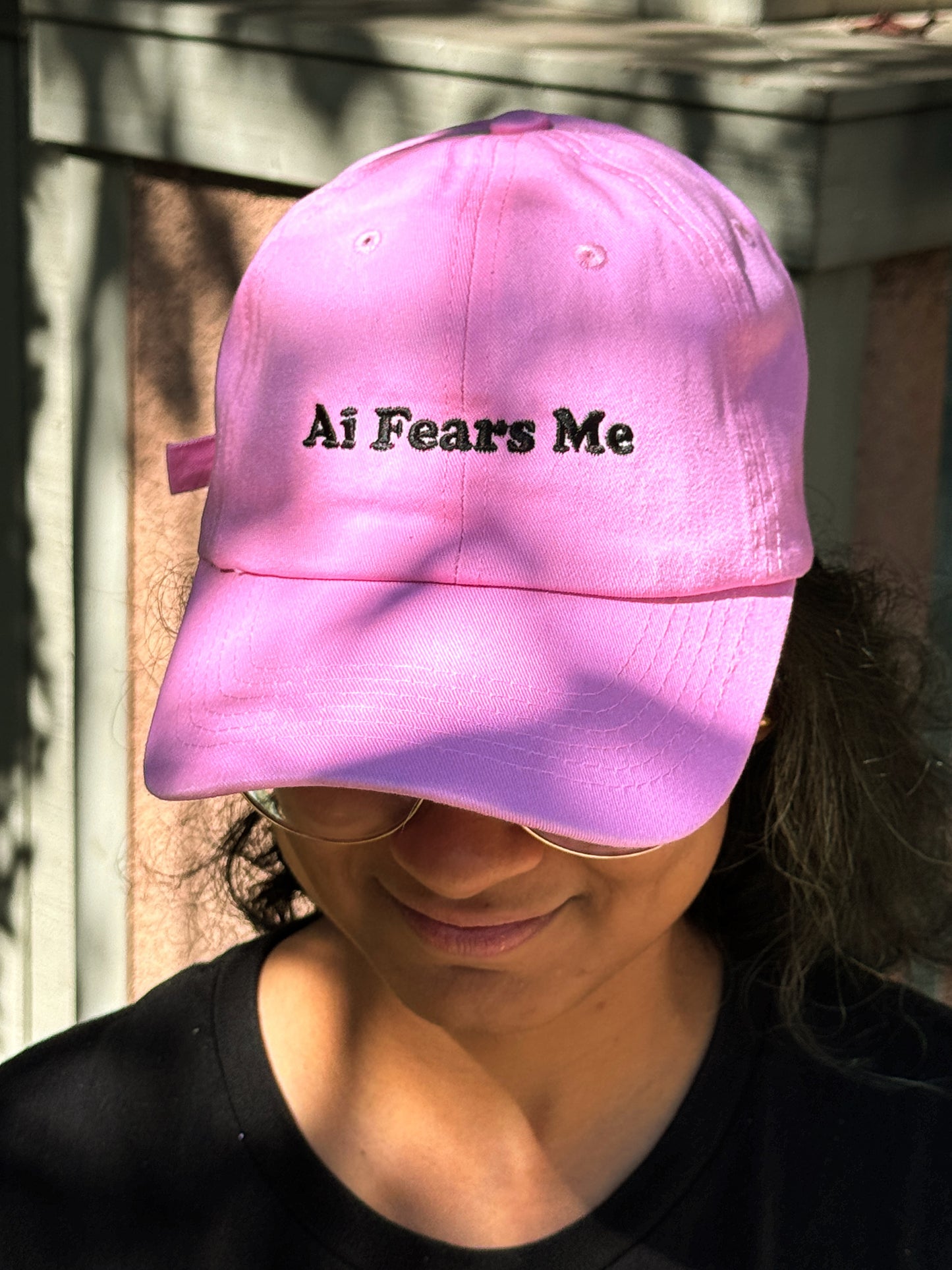 Person wearing a pink dad hat with the words 'AI Fears Me' embroidered in black on the front, standing outside in a shaded area.