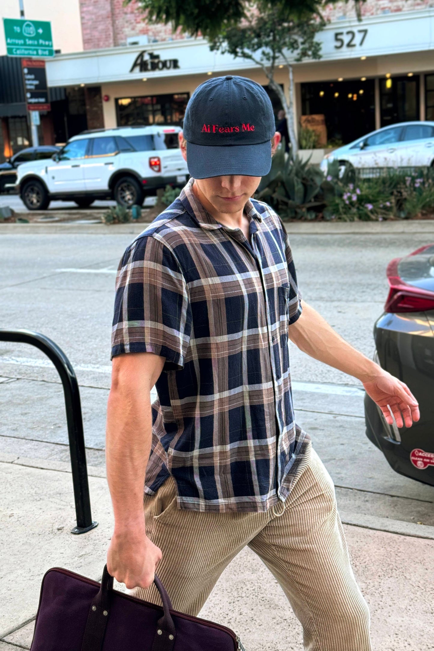 Person walking outdoors in a city, wearing a navy dad hat with the words 'AI Fears Me' embroidered in red on the front, paired with a plaid shirt and holding a bag.