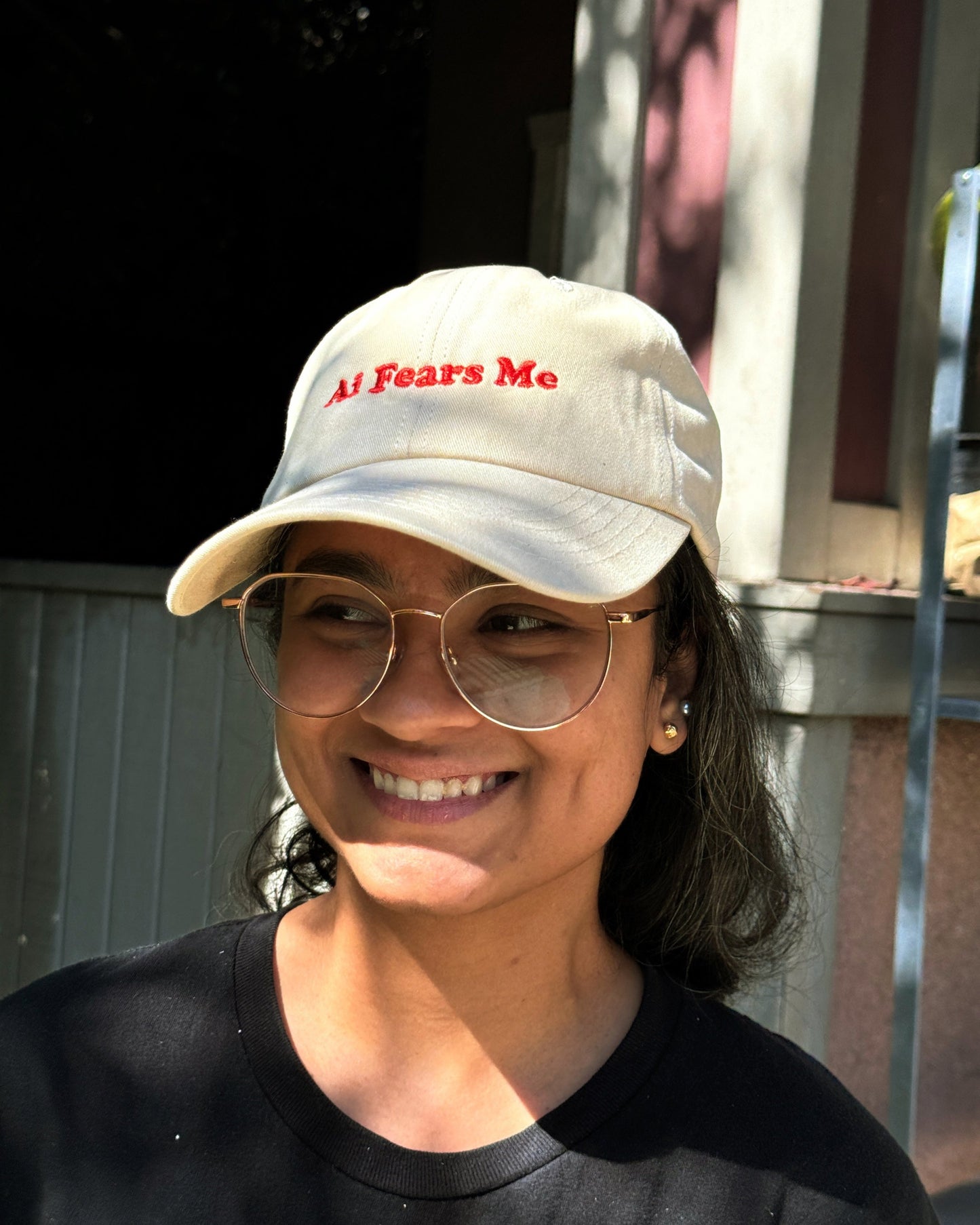 Person smiling while wearing a beige dad hat with the words 'AI Fears Me' embroidered in red on the front, standing outdoors in sunlight.