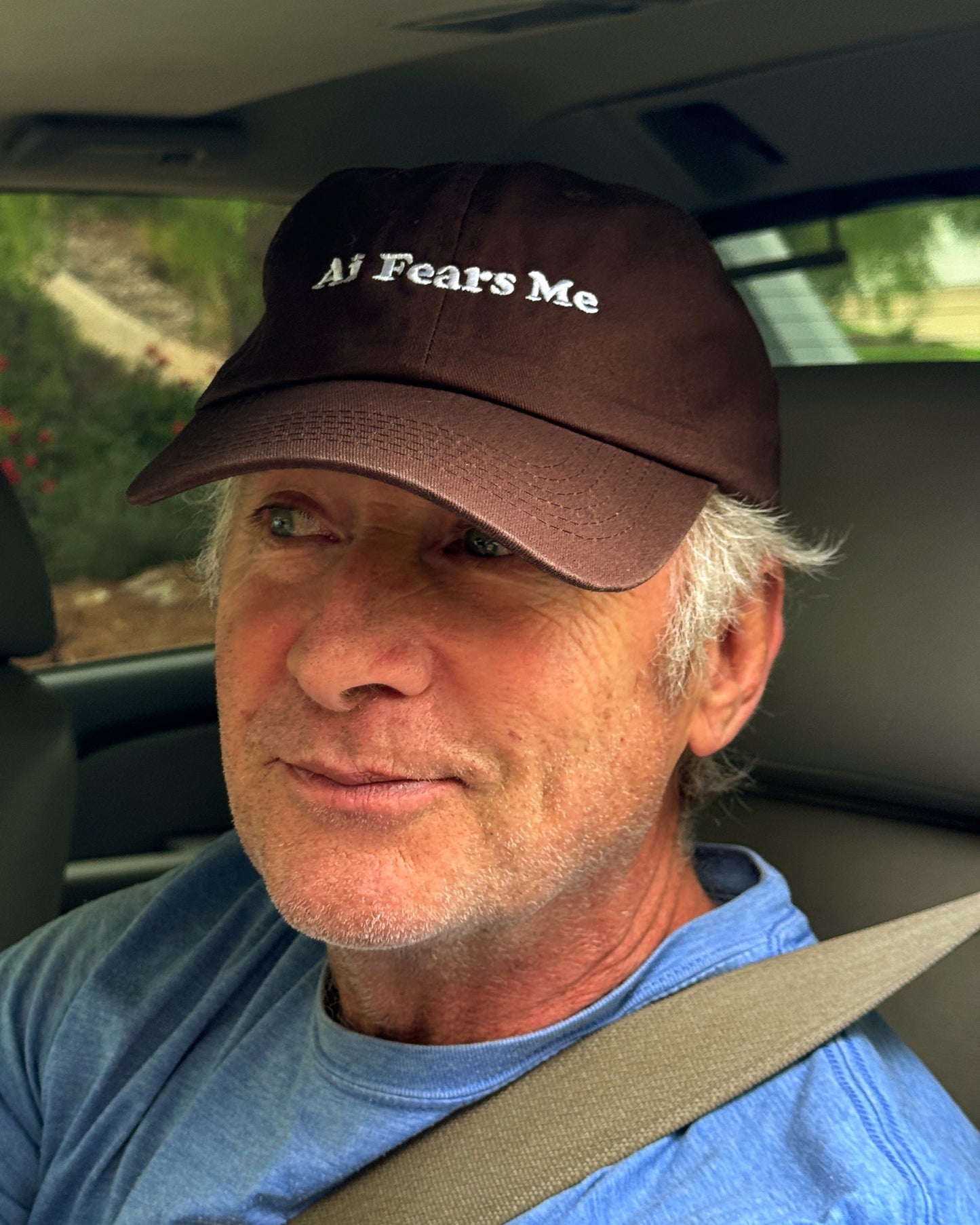 Person wearing a brown dad hat with the words 'AI Fears Me' embroidered in white on the front, sitting inside a car with a seatbelt fastened.