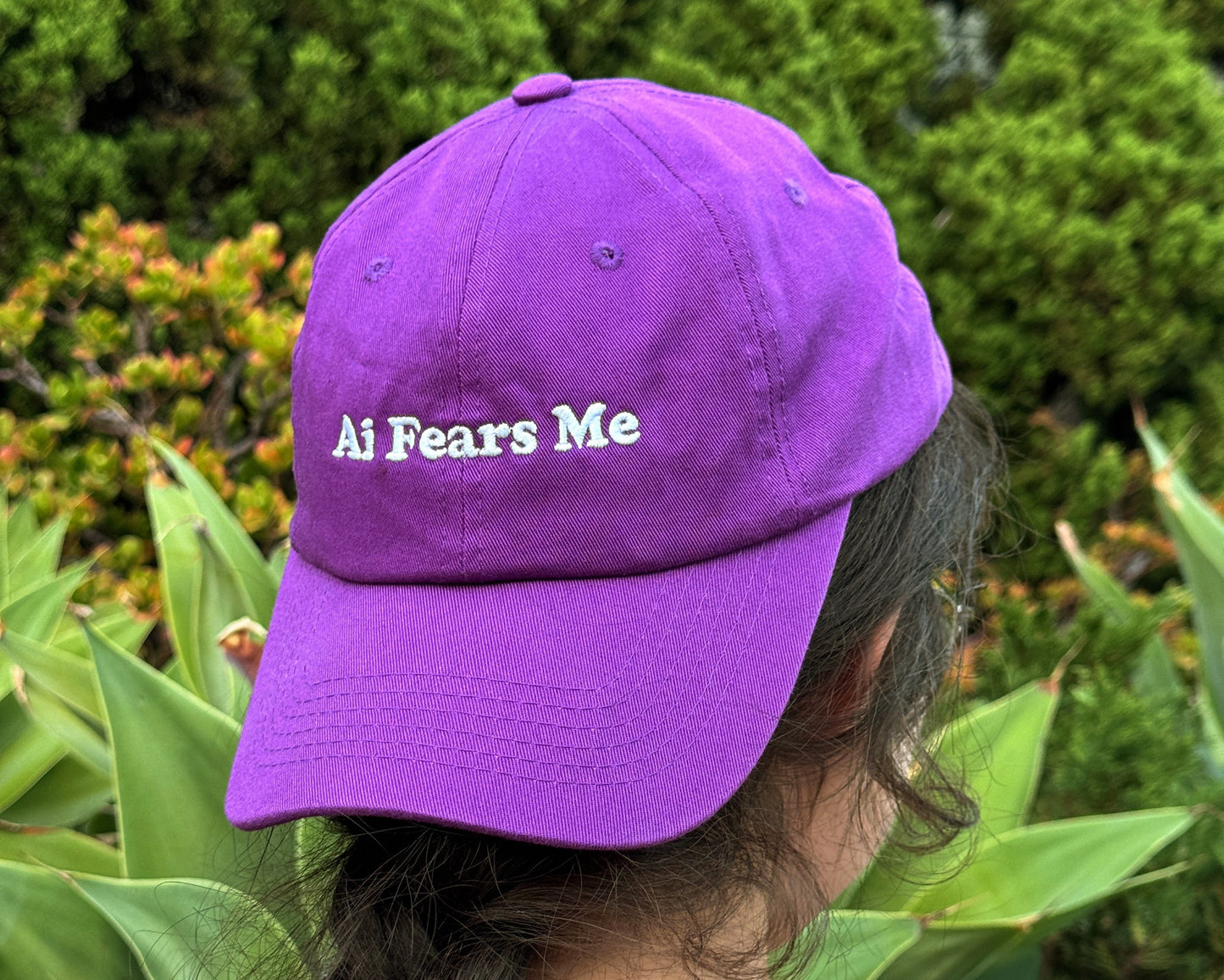 Person wearing a purple dad hat with the words 'AI Fears Me' embroidered in white on the front, seen from the back in an outdoor garden setting.